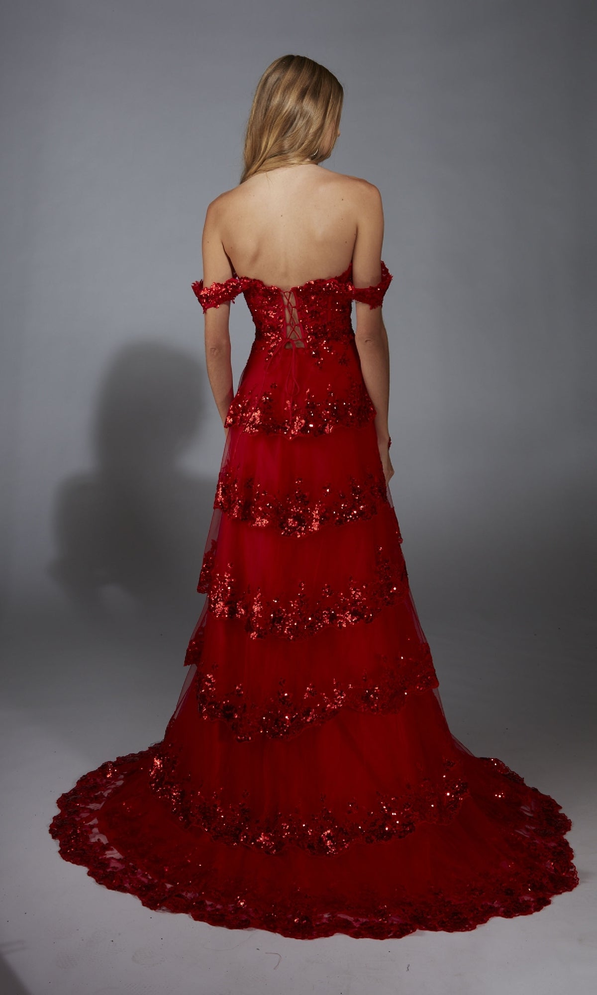 A woman with long blonde hair models the Aerie Couture 333, a tiered red sequin-embellished off-the-shoulder prom dress with a corset bodice, photographed from the back against a plain gray background.
